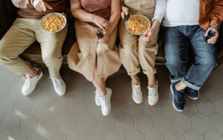 Na foto família sentada ao sofá comento pipoca e tomando refrigerante assistindo a um filme