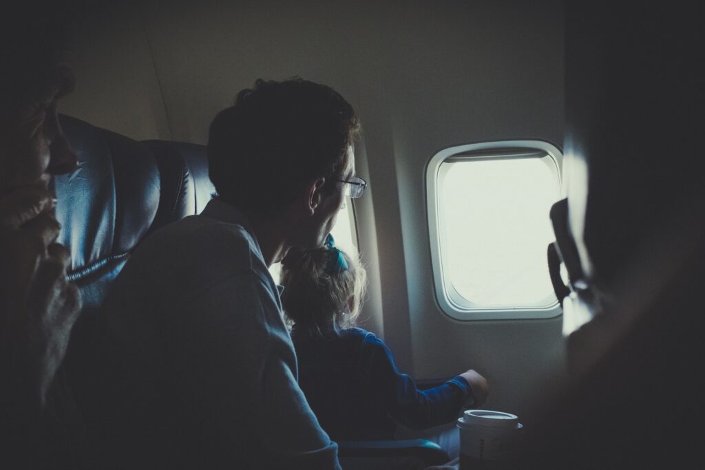 Na foto: pai viajando de avião com filho bebê em seu colo. Acento da janela. 