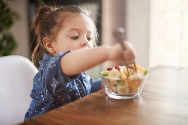 Na foto, menina pequena está sentada em frente a uma mesa esticando o braço para espetar frutas que encontram-se em um pote. Imagem/Reprodução: gpointstudio - Freepik