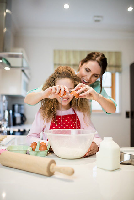 Na imagem, Mãe quebrando um ovo sobre uma tigela. Filha na frente olha contente para a receita que está sendo preparada. Imagem/Reprodução: peoplecreations - Freepik