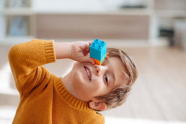 Na foto, menino com leve sorriso segurando peças de lego na frente de um dos olhos olhando em direção a quem o fotografa. Foto/Reprodução: Freepik